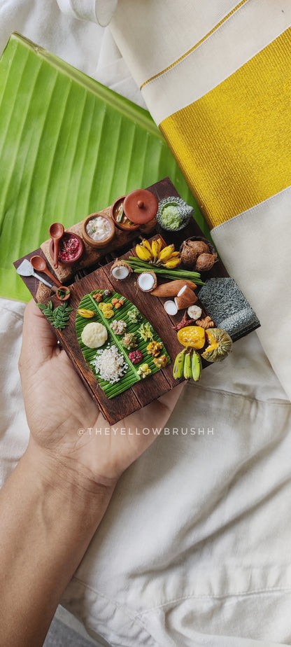 Miniature Onam festival feast display with banana leaves, fruits, coconuts, and serving bowls