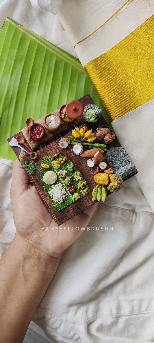 Miniature Onam festival feast display with banana leaves, fruits, coconuts, and serving bowls